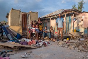 group of people young and old stand in front of a house destroyed, no walls, no windows