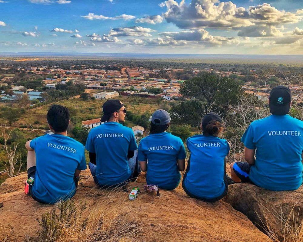 Thank you to volunteers: CMMB volunteers wear blue shirts while sitting on top of a mountain. They face the landscape and the back of their shirts read, 