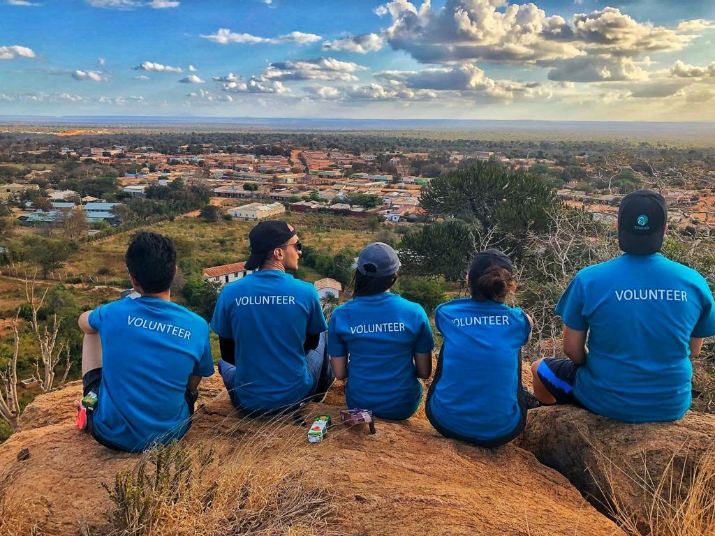 Thank you to volunteers: CMMB volunteers wear blue shirts while sitting on top of a mountain. They face the landscape and the back of their shirts read, "volunteer"