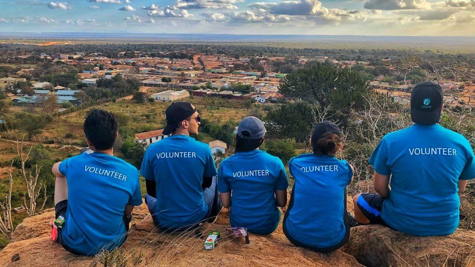 Thank you to volunteers: CMMB volunteers wear blue shirts while sitting on top of a mountain. They face the landscape and the back of their shirts read, 