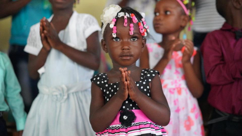 A young girl wearing a pink and black dress, stands with her hands in prayer.