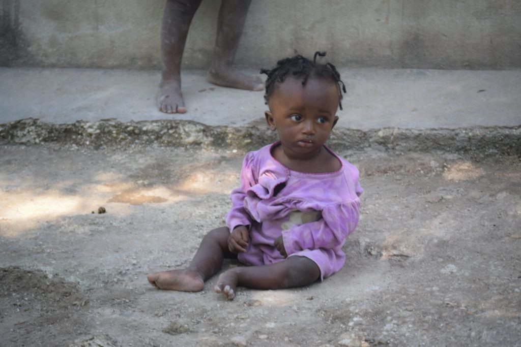 A young child sits on the gravel. He looks sad and weak.