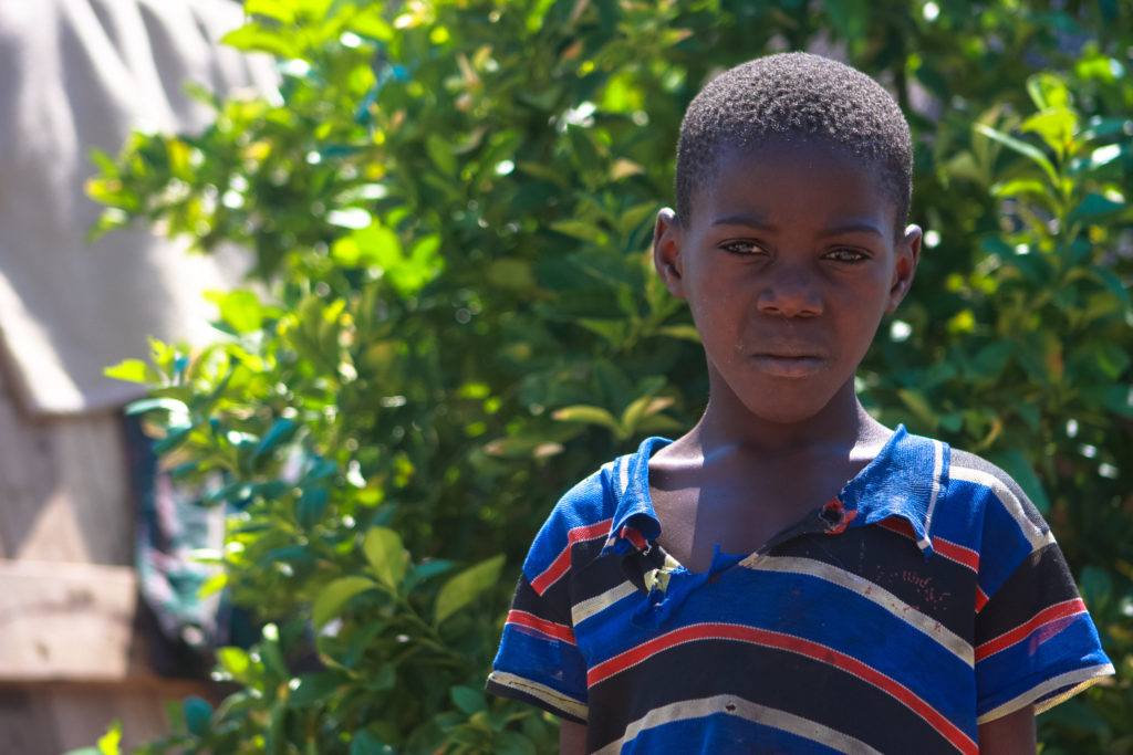 A young boy from Zambia stands in front of a green tree wearing a striped shirt