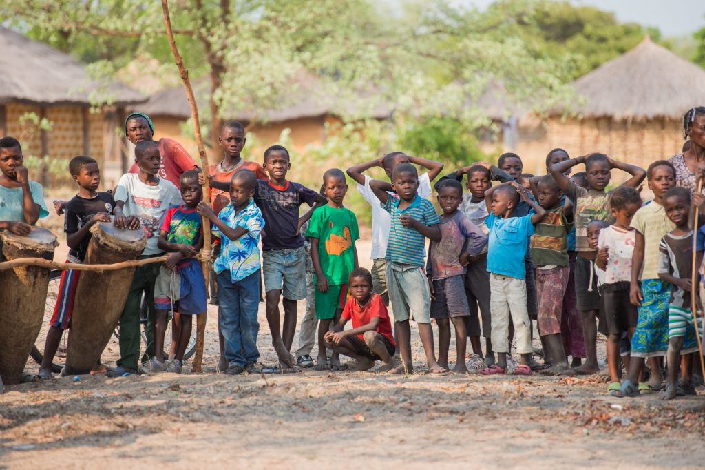 A group of children in Zambia standing together. In the background are homes and trees 