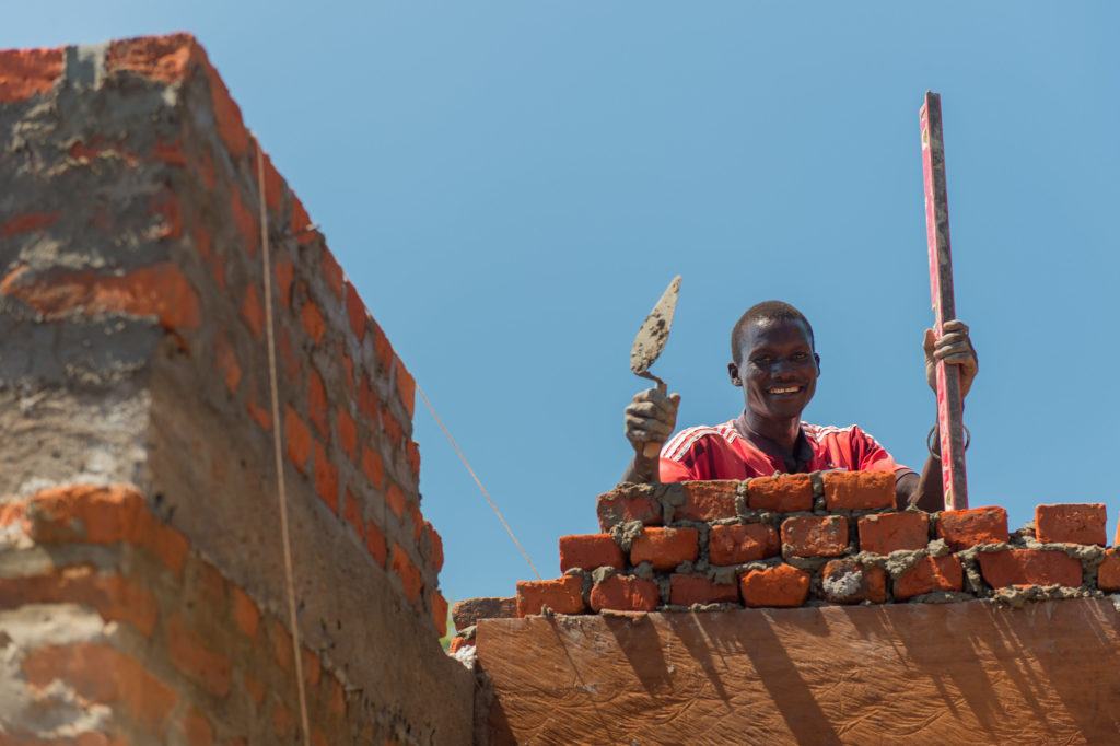 A member of the Hospital Construction Crew working. He holds tools in his hand as he smiles for the photo