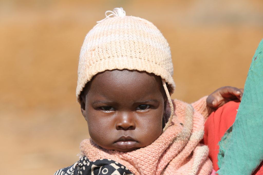 A young child with a pink hat is held in her mother's arms in Kenya. She is looking a little grumpy. 