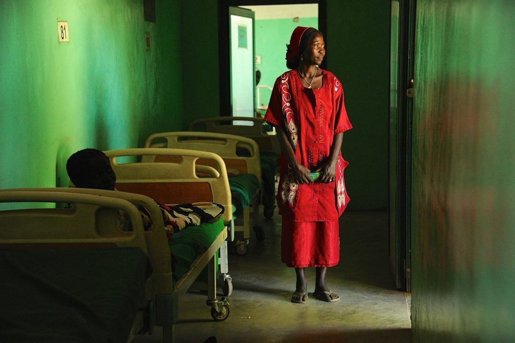 A women in a red outfit stands beside the bed of a loved one