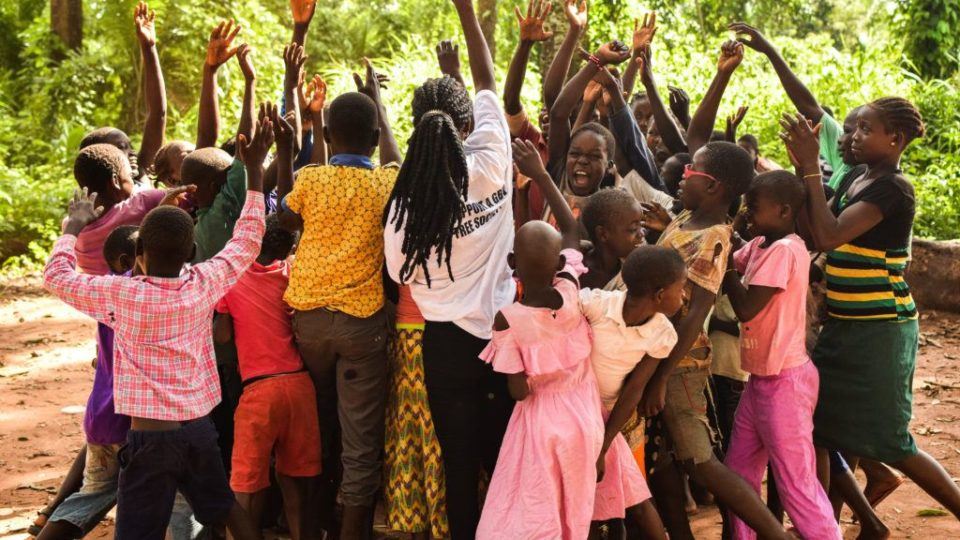 A group of South Sudanese children stand in a circle with an adult with hands raised. Everyone joins hands at the end of a CFS activity in South Sudan. We serve women and children living in these remote areas.
