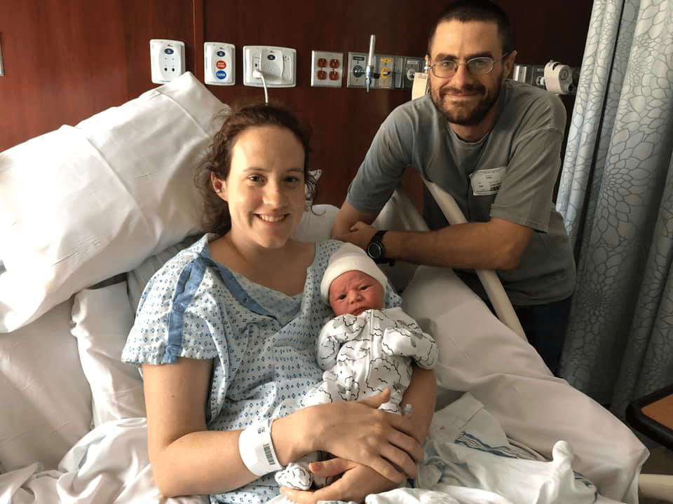 A new mom holds her newborn and her husband stands behind her hospital bed. This is the Rubino family. Sarah and Martin served at St. Therese Hospital in South Sudan.
