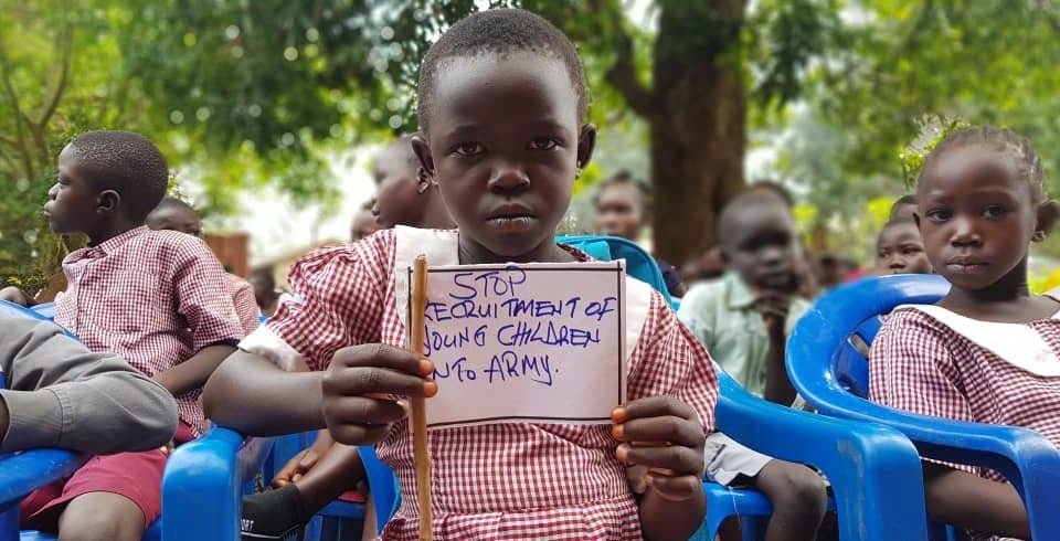 A young child holds a white flag that says: Stop the recruitment of children soldiers into the army