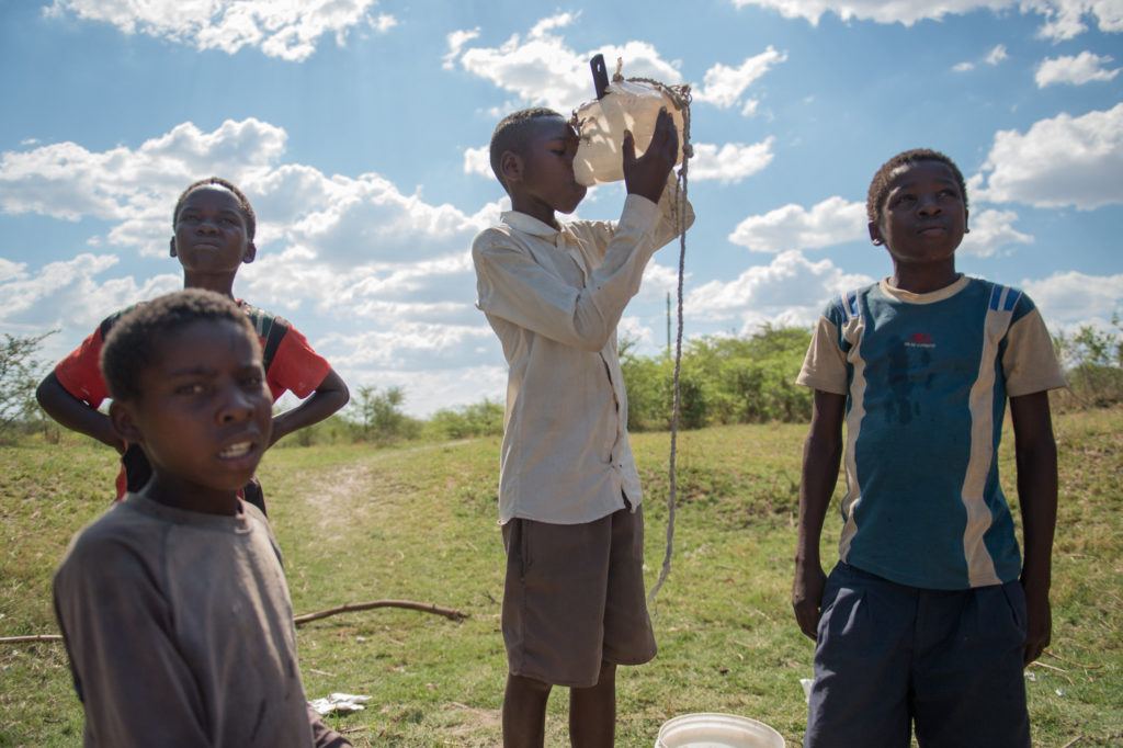 A boy drinks water from a jug. He stands outside in Zambia. other children stand around him