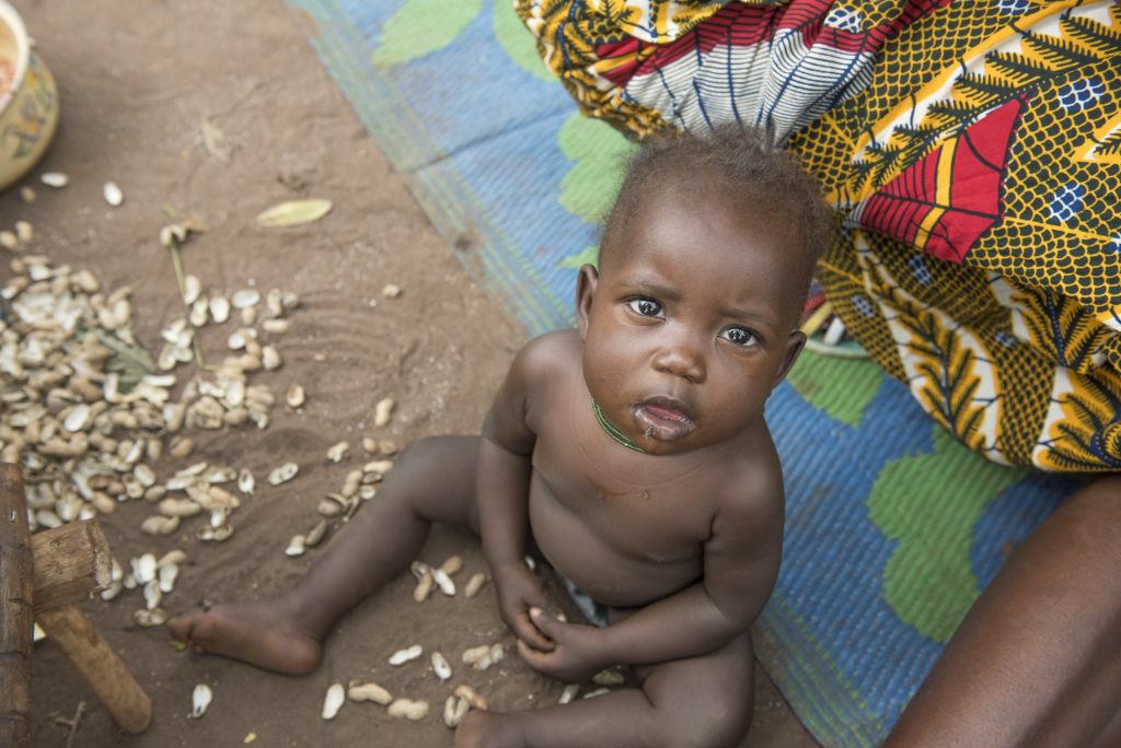 A child sits on the ground looking upwards. He sits on the dirt with a blue blanket behind him 