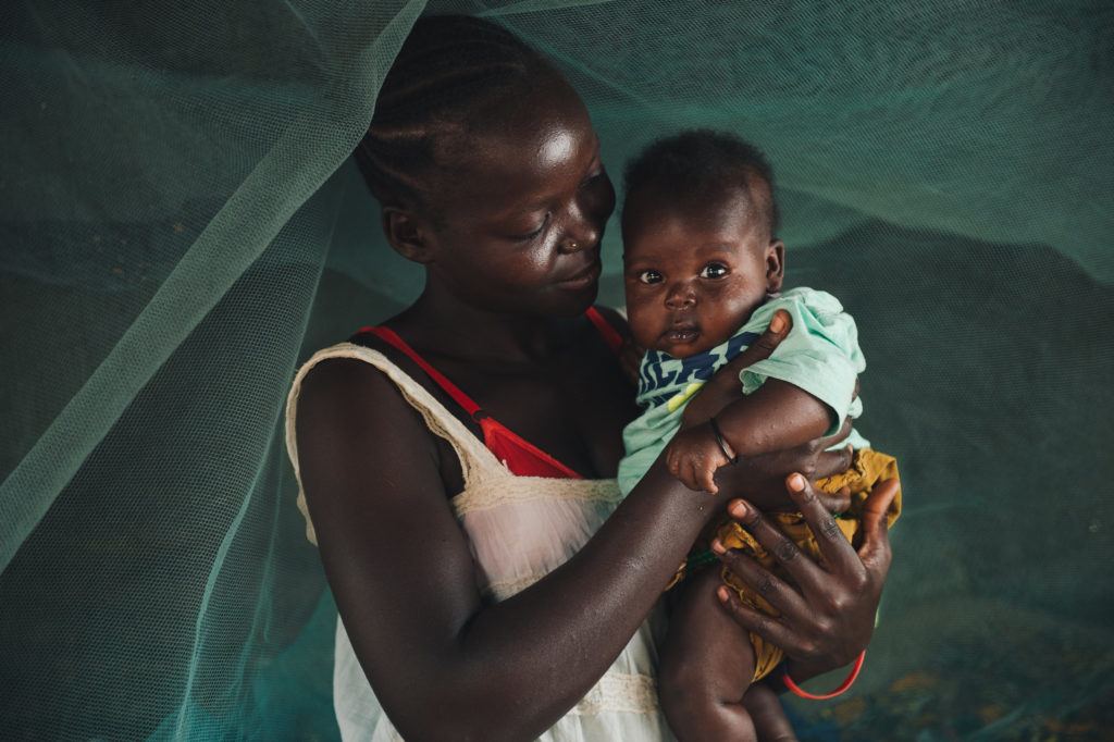 A young mother holds her child. They stand under a mosquito net. She is among the rural women that we celebrate on international day of rural women.