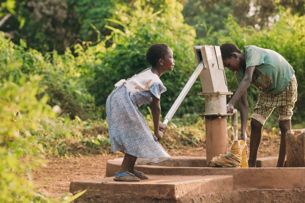 A young girl pumps water from a well in South Sudan. There is greenery around her and she is joined by another child. 