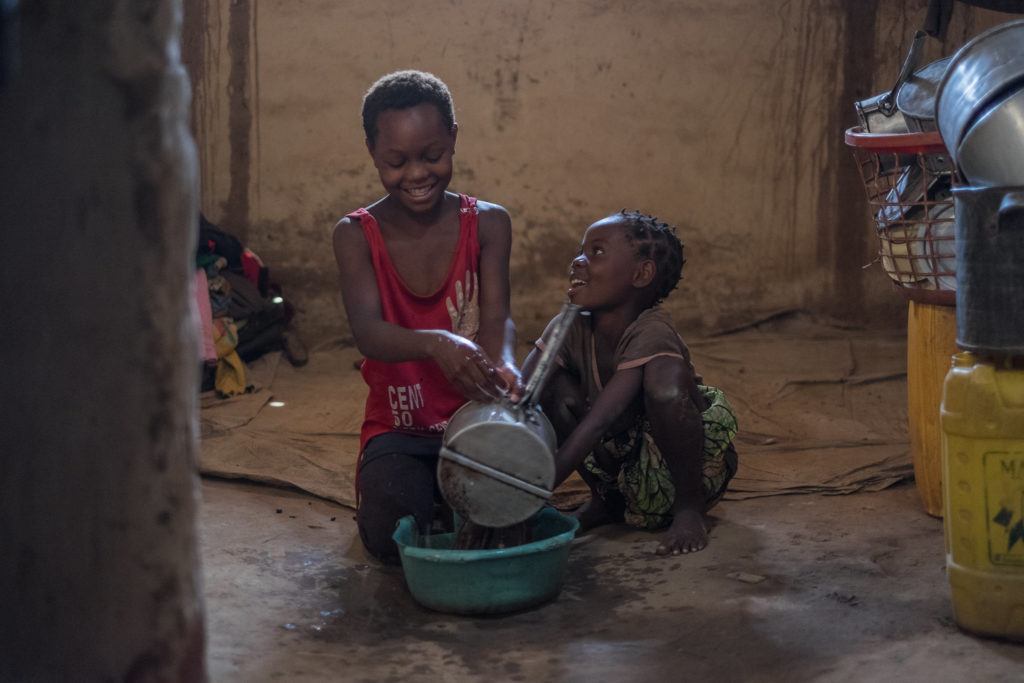 Two children sit in front of a water bucket on the floor of their home. 