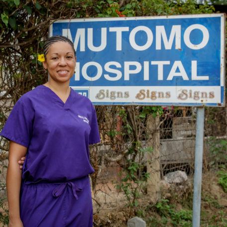 Community-Healthcare-Worker-Mutomo-Hospital-Sign A community health care worker standing in front of Mutomo Hospital sign.