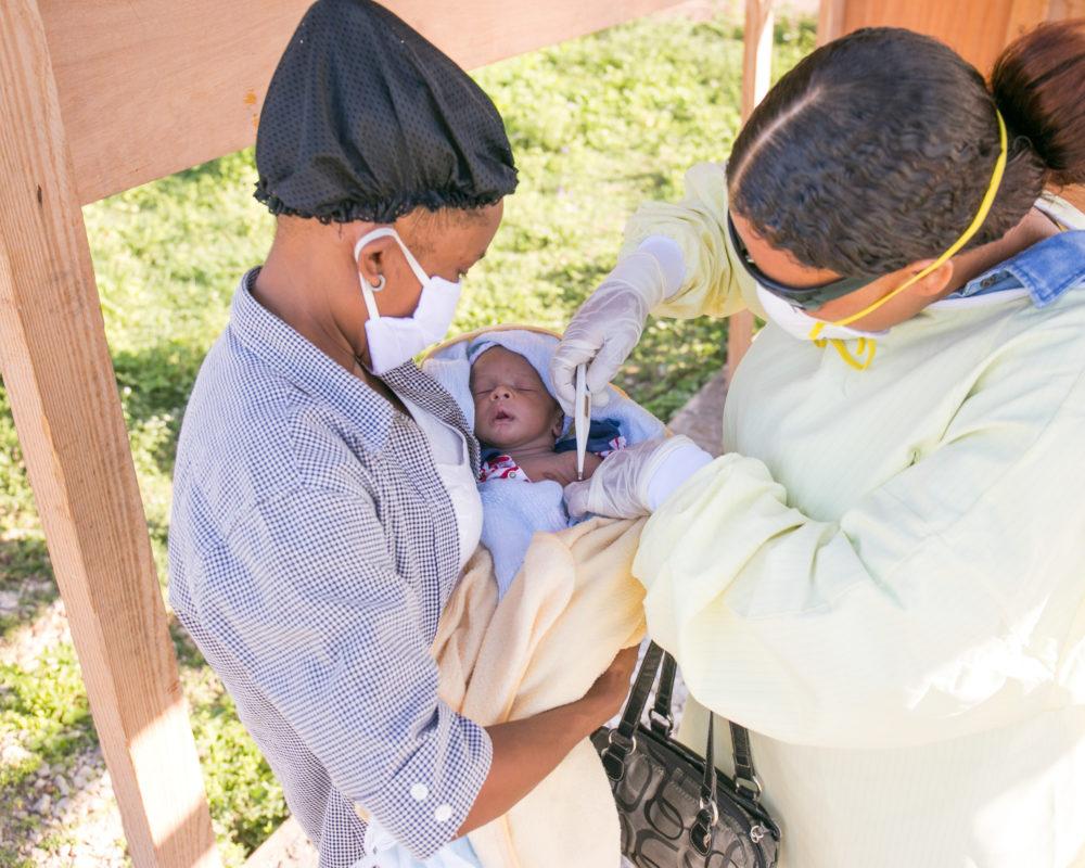 A mother holding her infant child as a community healthcare worker in Haiti assist for COVID-19 in Haiti in July 2002.