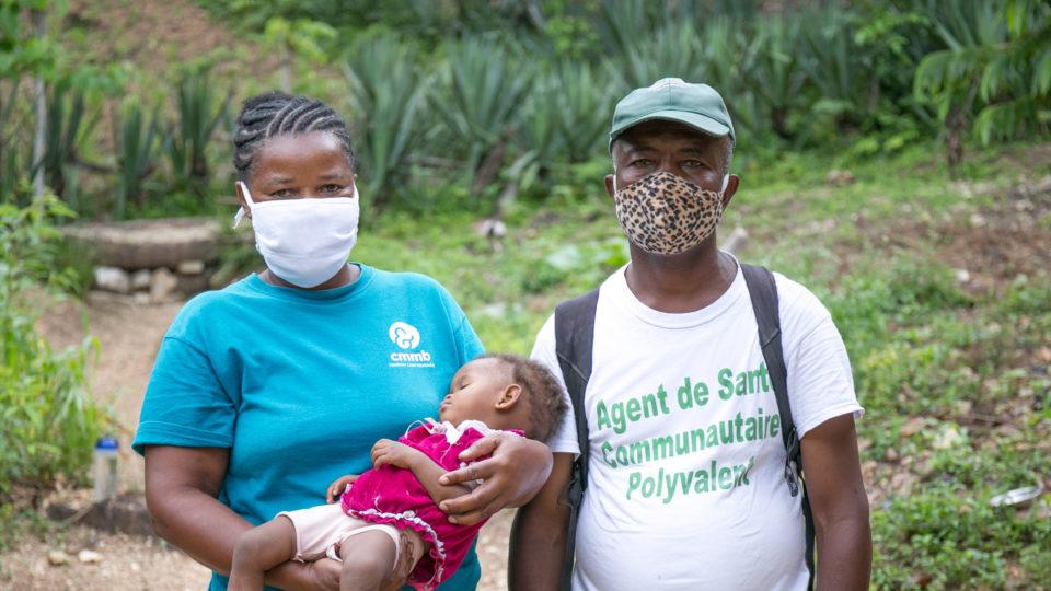 Community health worker wearing masks and holding a child in Haiti during COVID-19.