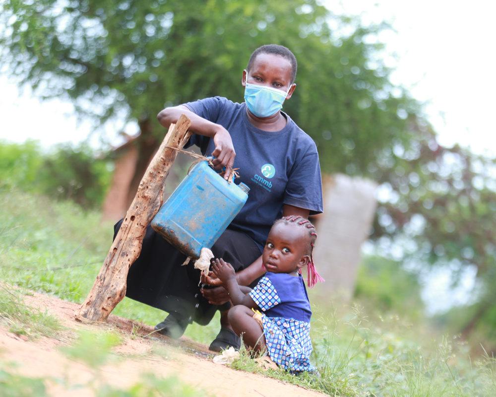 A community health worker wearing a face mask with a young girl at a tippy tap for clean water in Kenya in 2021.