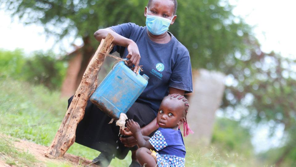 A community health worker wearing a face mask with a young girl at a tippy tap for clean water in Kenya in 2021.