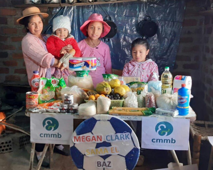A mother with her young children stand before a survival care package for COVID-19 provided by CMMB in Peru in 2021.
