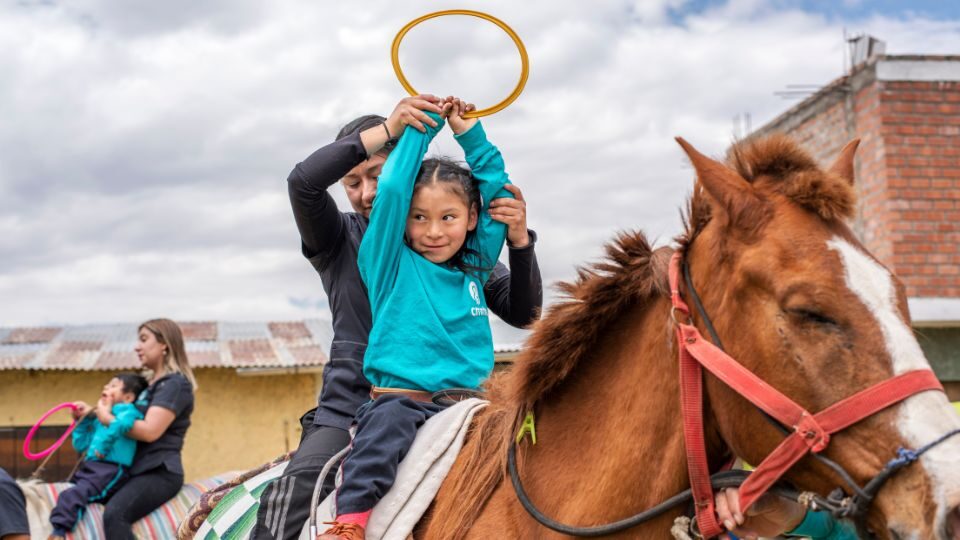 Rosalinda sits astride a horse holding a plastic ring over her head during CMMB-supported equine therapy
